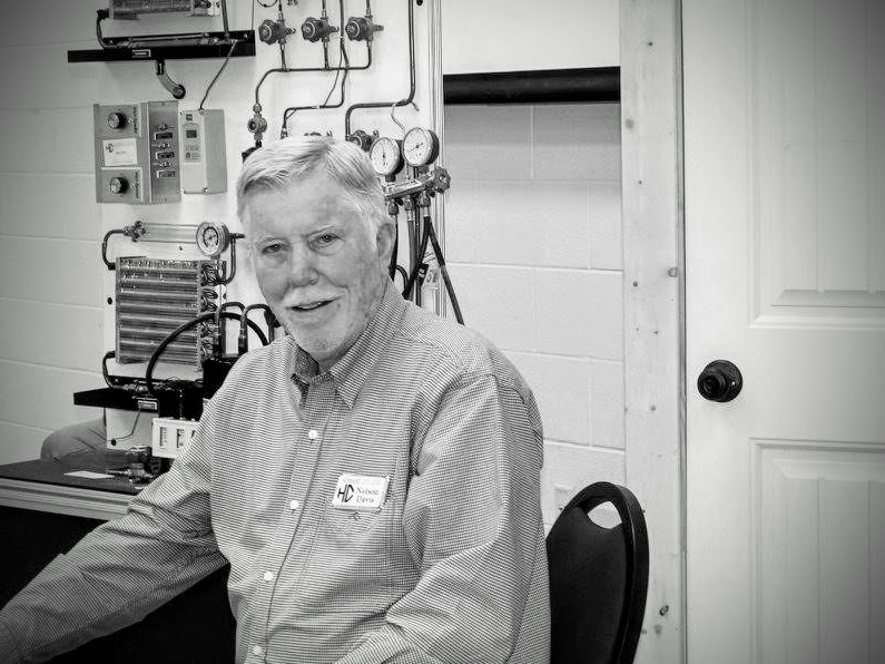 Greyscale image of Nelson Davis sitting in office, with a display of gauges and air conditioning parts in the background